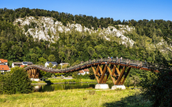 Holzbrücke über einen Fluss mit bewaldetem Hügel im Hintergrund.