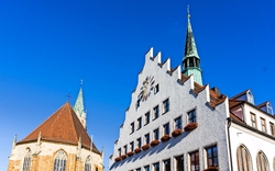 Ein historisches Gebäude mit Giebeldach und Uhr auf der Fassade neben einer Kirche mit zwei Türmen unter einem klaren blauen Himmel.