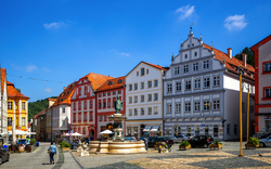 Historischer Marktplatz mit bunten, kleinen Stadthäusern im Barockstil und einem zentralen Brunnen mit einer Statue. Einige Menschen spazieren auf dem Kopfsteinpflaster bei sonnigem Wetter.