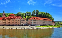 Ein großes Klostergebäude mit rotem Dach am Ufer eines ruhigen Flusses, umgeben von grünen Wäldern unter einem klaren blauen Himmel.