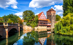 Ein malerisches Flussufer in Nürnberg, Deutschland, mit traditioneller Fachwerkarchitektur und einem historischen Steingebäude. Eine Brücke führt über den Fluss, und das Wasser spiegelt die umliegenden Bäume und den blauen Himmel mit weißen Wolken wider.