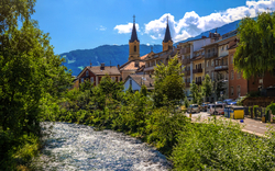Fluss fließt durch malerische Stadt mit Kirchtürmen, Bäumen und Bergen im Hintergrund.