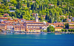 Stadtansicht am See mit italienischer Architektur und grünen Hügeln im Hintergrund.