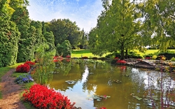 Ein gepflegter Garten mit einem Teich, umgeben von üppigen grünen Bäumen und bunten Blumenbeeten.