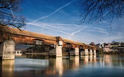 Holzbrücke über einen Fluss mit blauem Himmel und Dorf im Hintergrund