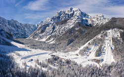 Schneebedeckte Berglandschaft mit Skipiste und Bäumen unter blauem Himmel.