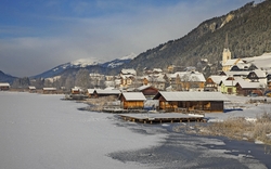Verschneites Dorf am See mit Bergen im Hintergrund bei sonnigem Wetter.