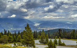 Berglandschaft mit See, Wald und Wolkenhimmel im Hintergrund.
