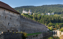 Burg auf einem bewaldeten Hügel mit Stadtmauer im Vordergrund