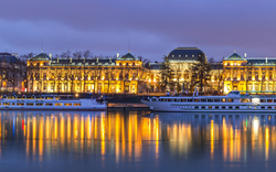 Panorama einer beleuchteten Stadtansicht mit Fluss und Gebäuden bei Nacht.