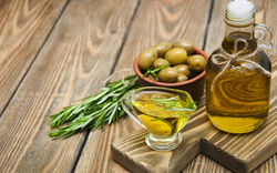 Olivenöl in Flasche neben Oliven und Rosmarinzweigen auf Holztisch.