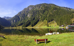 Bänke am Ufer eines Bergsees mit Wald und Berghütte im Hintergrund.