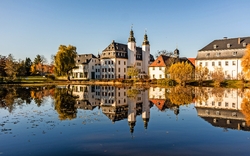 Schloss und historische Gebäude spiegeln sich in einem ruhigen See an einem sonnigen Herbsttag, umgeben von buntem Herbstlaub.