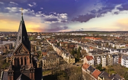 Luftaufnahme einer Stadtlandschaft bei Sonnenuntergang, mit einer Kirche im Vordergrund und einer Ansammlung von Gebäuden und Bäumen im Hintergrund.