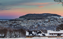 Panoramablick auf eine verschneite Stadtlandschaft bei Sonnenuntergang, mit einem Hügel im Hintergrund und Häusern im Vordergrund, die von schneebedeckten Bäumen umgeben sind.