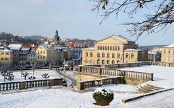 Blick auf eine winterliche Stadtlandschaft mit schneebedeckten Gebäuden und Straßen, im Vordergrund eine steinerne Balustrade, im Hintergrund sind historische Gebäude und ein Kirchturm sichtbar.
