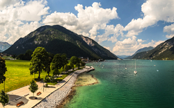 Blick auf eine Seepromenade, grüne Wiesen und Berge bei sonnigem Wetter.