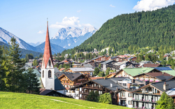 Kirchturm in einem Alpenort vor bewaldeten Bergen und klarer Himmel.