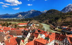 Stadtansicht mit roten Dächern, Fluss und Bergen im Hintergrund unter blauem Himmel.