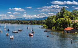 Segelboote auf einem See mit bewaldeten Ufern und Bergen im Hintergrund.