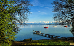 See mit Steg, Booten und Bergen im Hintergrund an einem sonnigen Tag.