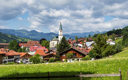 Alpenlandschaft mit Kirche und Dorf, grüne Wiesen, Berge im Hintergrund.