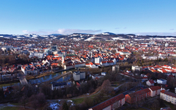 Luftaufnahme einer Stadt mit Fluss und umliegendem Hügelland im Hintergrund.