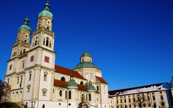 Barocke Kirche mit Doppeltürmen und benachbartem Gebäude vor blauem Himmel.