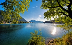 Bergsee mit umliegenden Bäumen bei Sonnenschein und blauem Himmel.