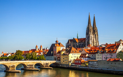 Stadtansicht mit Brücke und Kathedrale an einem Fluss bei Sonnenschein.