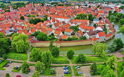 Luftaufnahme einer historischen Stadt mit roten Dächern und Stadtmauer, umgeben von Grünflächen.