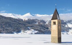 Verschneiter Kirchturm im Reschensee vor schneebedeckten Bergen.