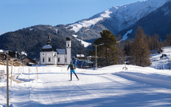Langläufer auf verschneiter Strecke vor Alpenkirche und Berglandschaft.