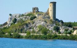 Turmruine auf einem felsigen Hügel an einem Flussufer mit Bäumen und blauem Wasser.